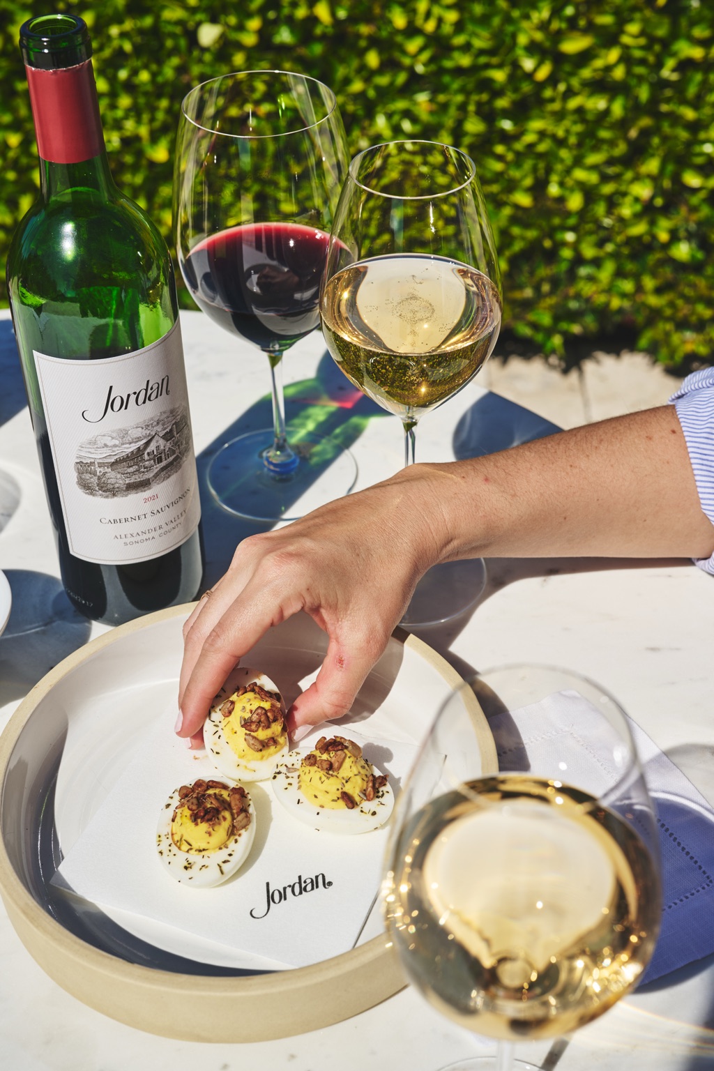 Hand reaching for deviled eggs beside a bottle of Jordan Alexander Valley cabernet sauvignon on a sunlit terrace