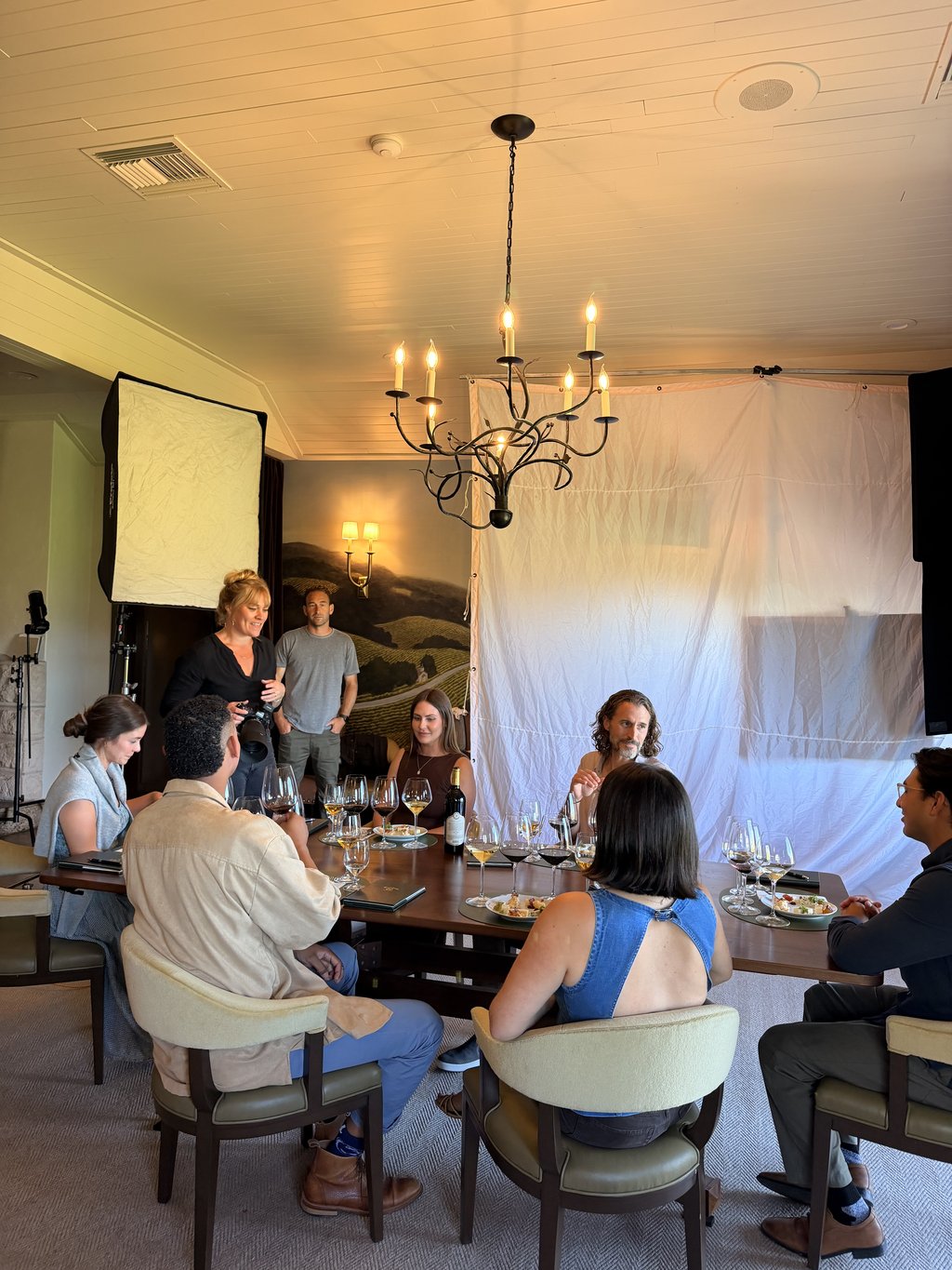 Dawn Heumann directs a wine dinner scene with talent seated around the table, softbox and muslin backdrop behind the chandelier