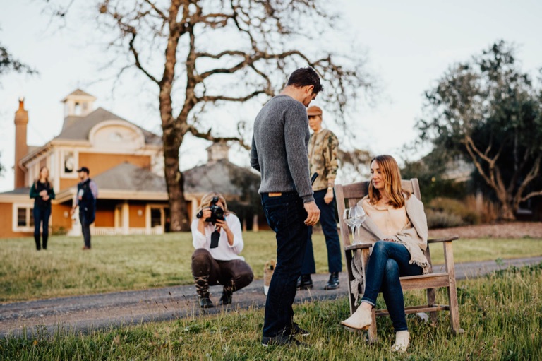 A photographer crouches to shoot talent seated on a bench during an on-location portrait session at an estate
