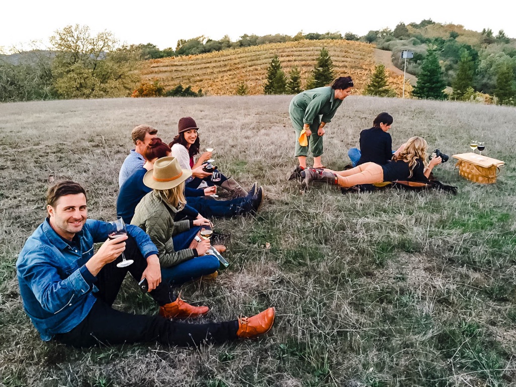 Crew and friends with wine glasses settled on a golden hillside above vineyard rows at dusk