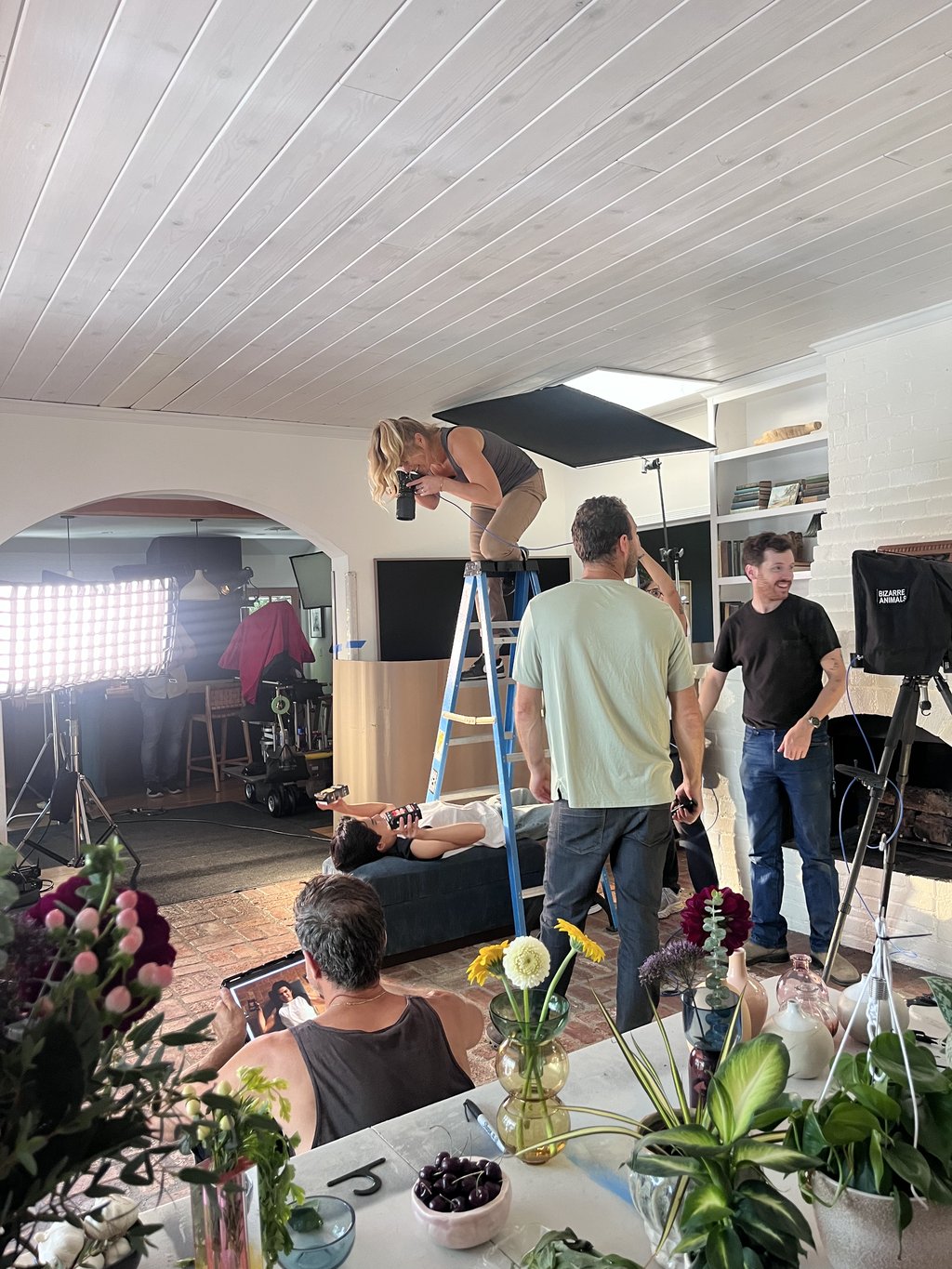 Dawn Heumann shoots down from a ladder at talent on a daybed while the crew sets lighting and a client reviews frames on a tablet