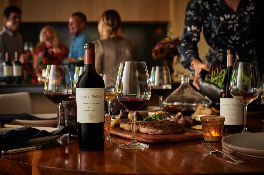 Lambert Bridge cabernet sauvignon at a walnut dinner table with steak board while friends gather behind