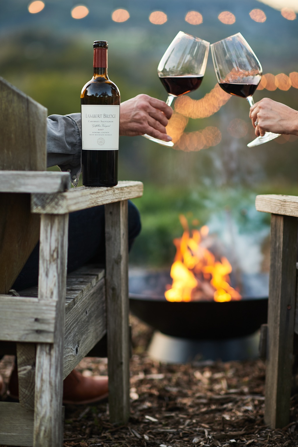 Couple toasting red wine over a fire pit at dusk, Lambert Bridge cabernet resting on the chair arm