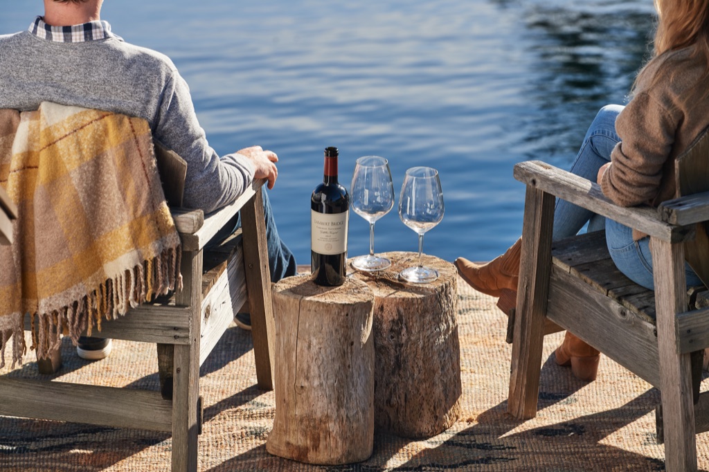 Couple in weathered Adirondack chairs at the water's edge with Lambert Bridge cabernet on a driftwood stump, photographed by Dawn Heumann