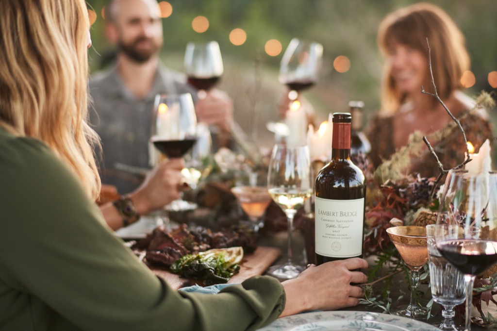 Woman presenting Lambert Bridge cabernet at an outdoor dinner party as friends raise glasses under string lights