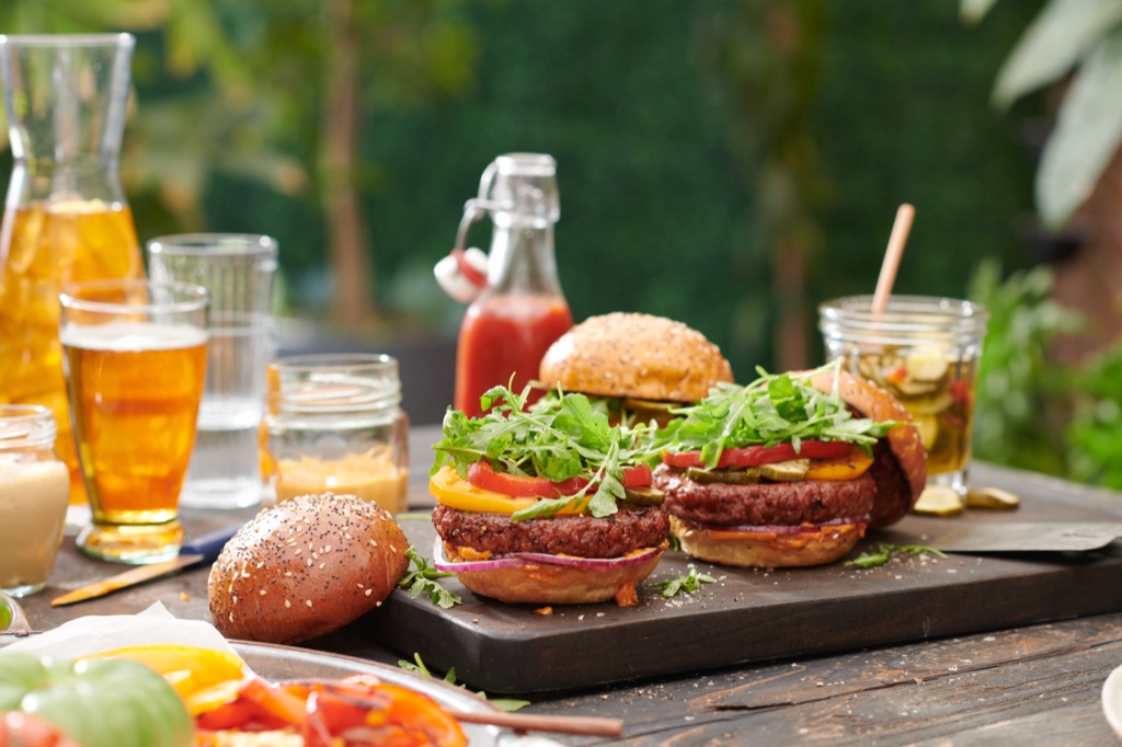 Burgers piled with arugula on a dark board with beer, pickles and hot sauce in the garden