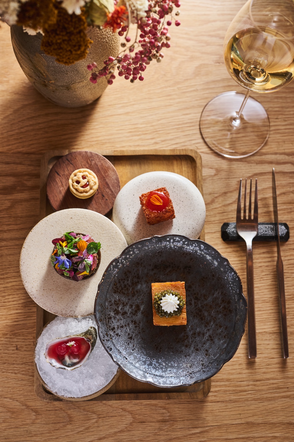 Overhead of a tasting menu canapé tray with a caviar pastry, an oyster on ice and white wine