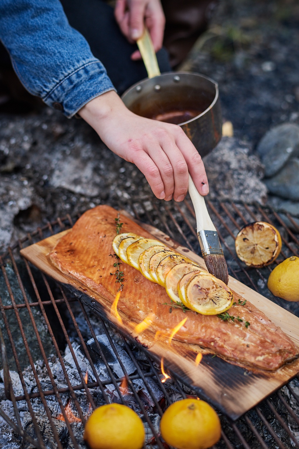 Brushing glaze onto cedar-plank salmon with charred lemons over a beach campfire