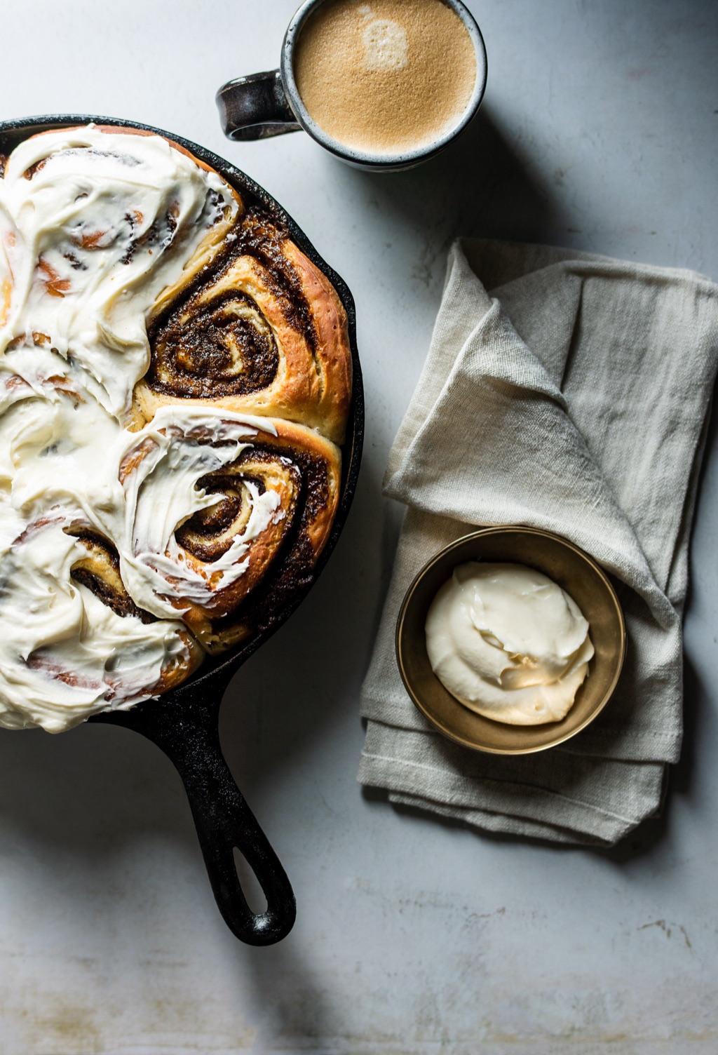 Overhead of frosted cinnamon rolls in a cast iron skillet with a latte and bowl of icing