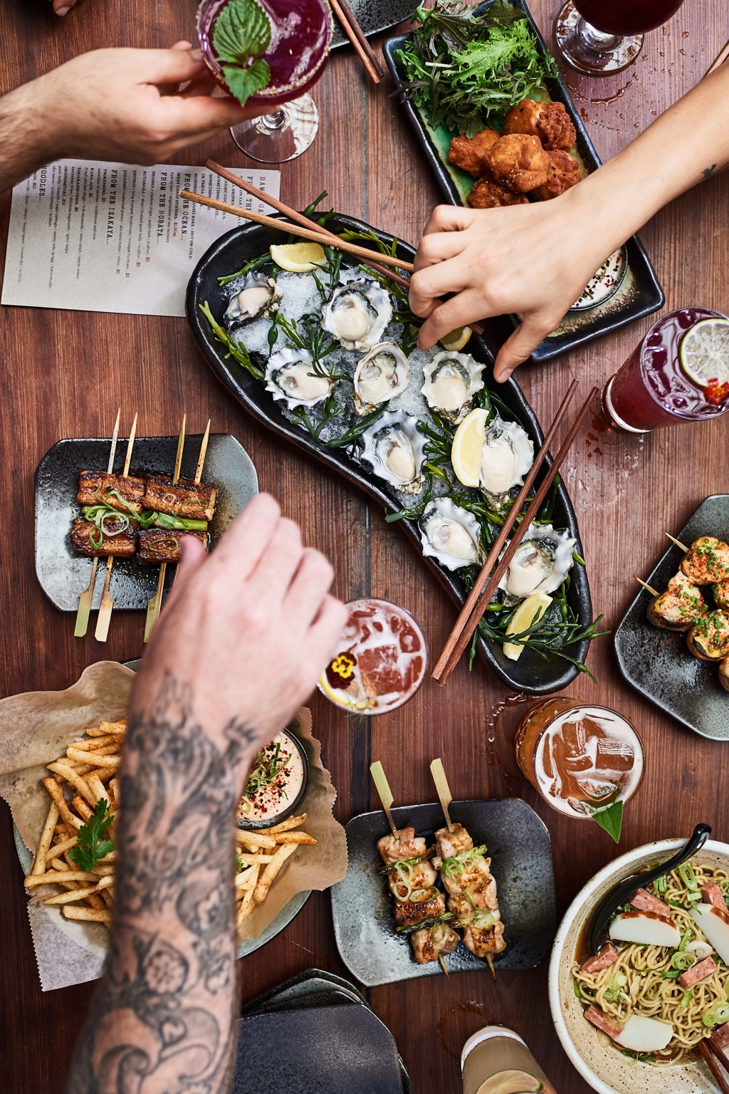 Overhead of friends reaching over an izakaya spread of oysters, skewers and cocktails