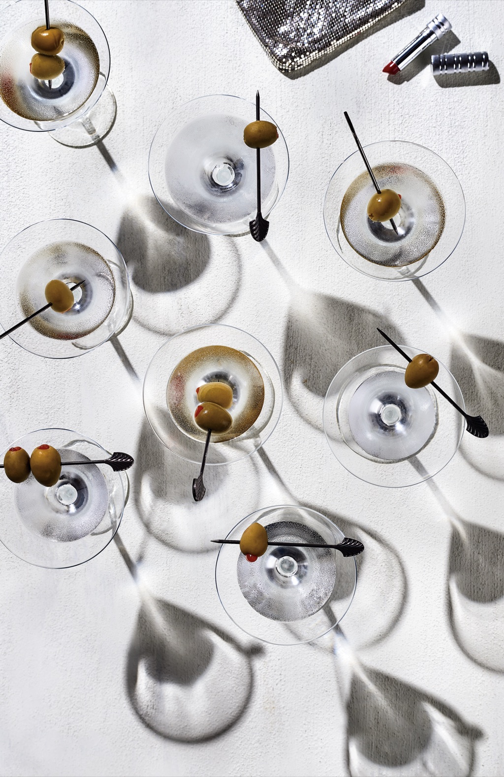 Overhead of dry martinis with olive picks casting hard shadows beside a sequin clutch and red lipstick
