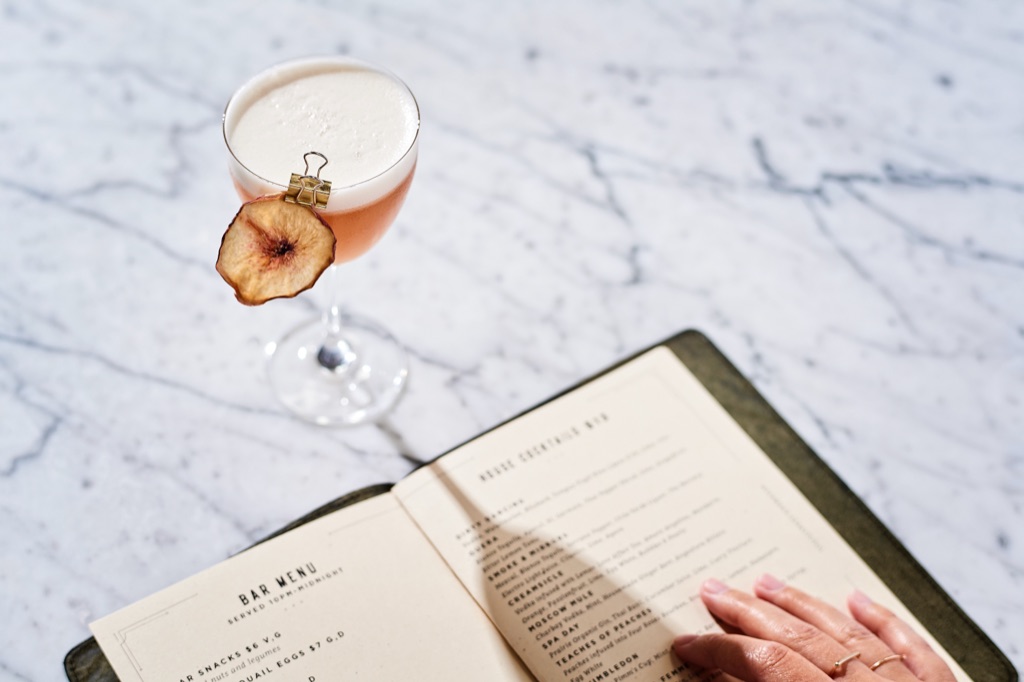 Peach sour with a dried-fruit garnish beside a hand browsing the bar menu on marble