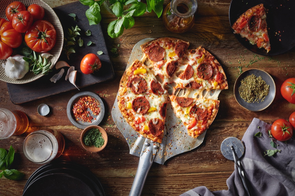 Overhead of pepperoni pizza on a metal peel with beer, basil and chili flakes on dark wood