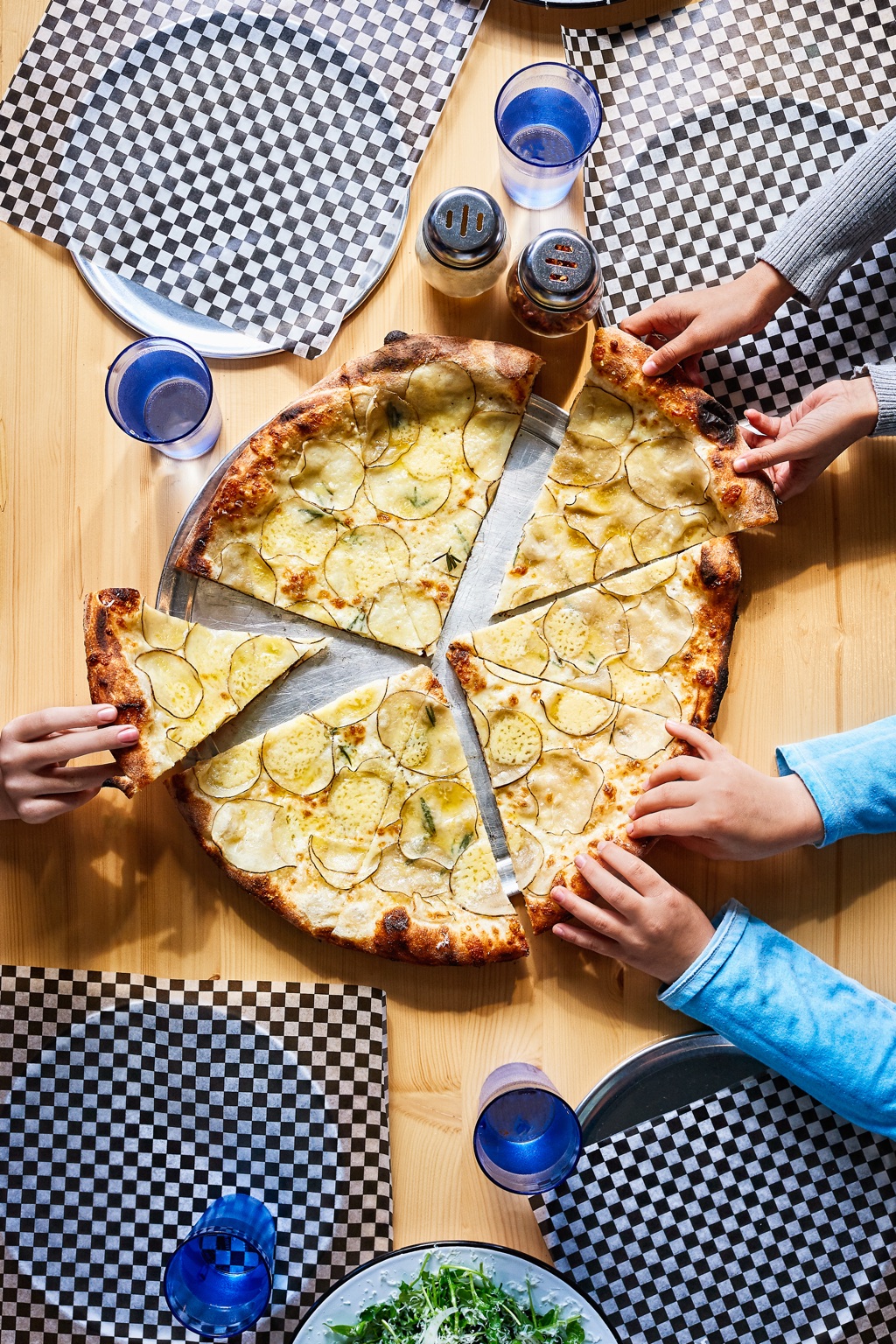 Overhead of kids' hands grabbing slices of rosemary potato pizza on checkered paper