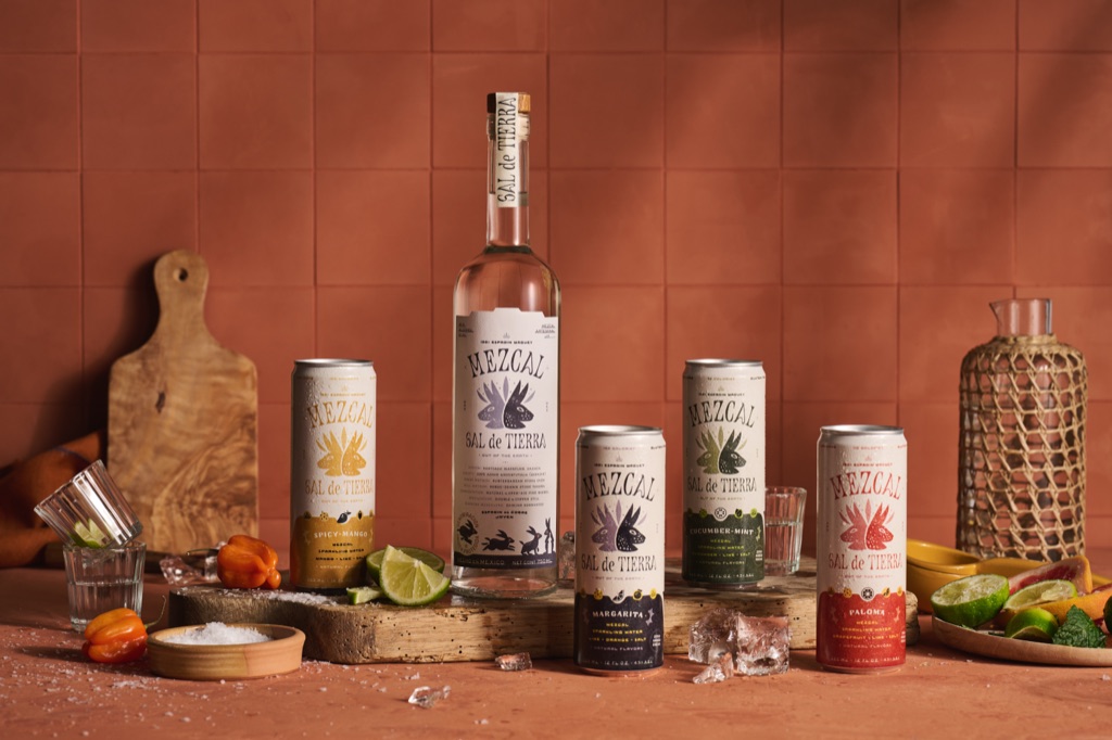Sal de Tierra mezcal bottle and four cocktail can flavors against terracotta tile