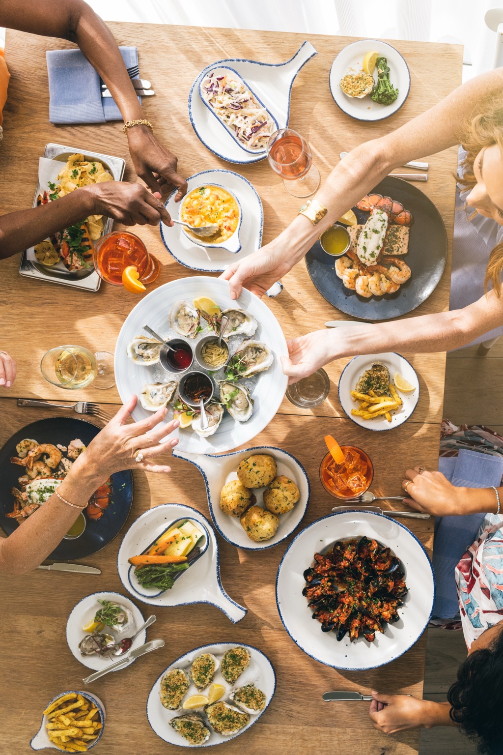 Overhead of friends passing an oyster platter over lobster, mussels and spritzes at a seafood restaurant