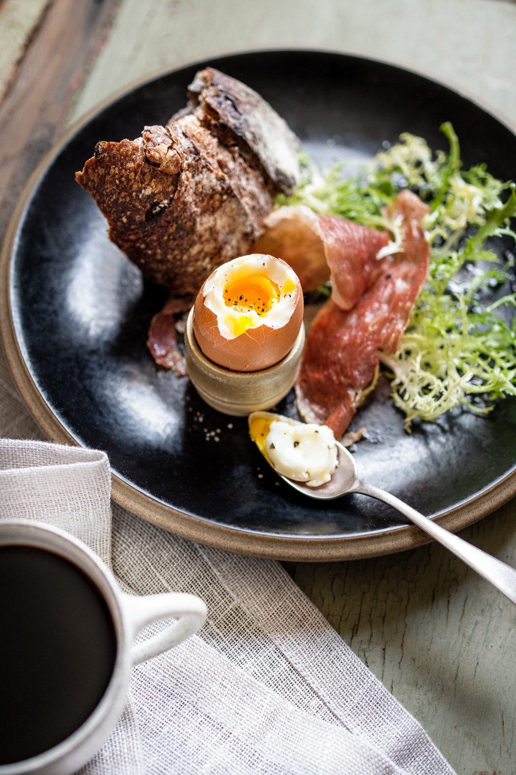 Soft-boiled egg in a ceramic cup with prosciutto, frisée and torn bread on a black plate