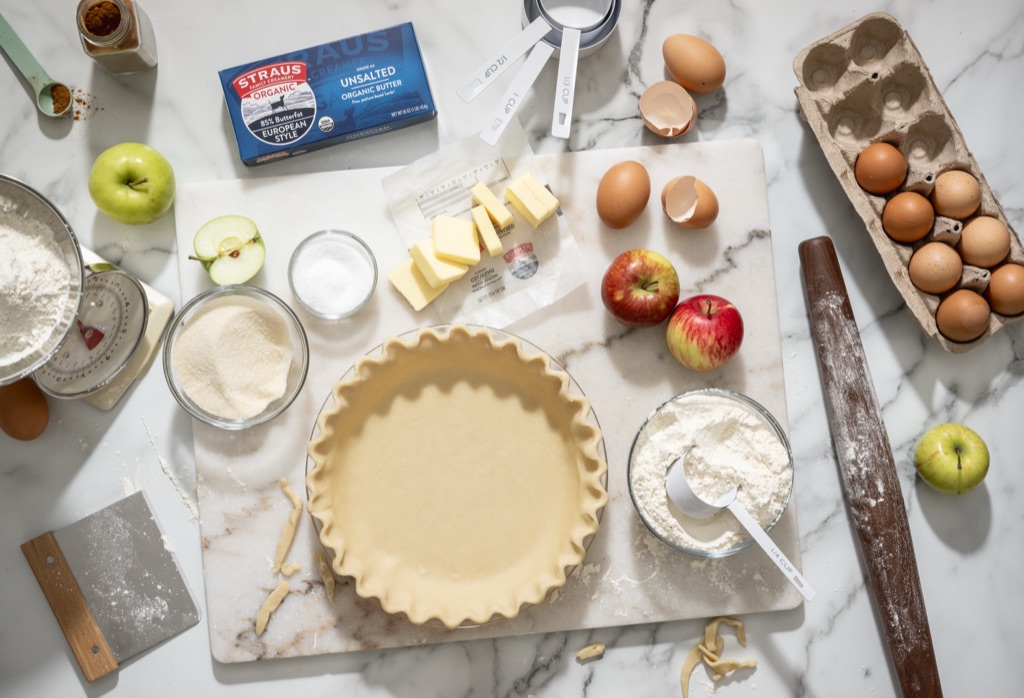 Overhead of apple pie baking with Straus organic butter, a crimped crust, apples and brown eggs on marble