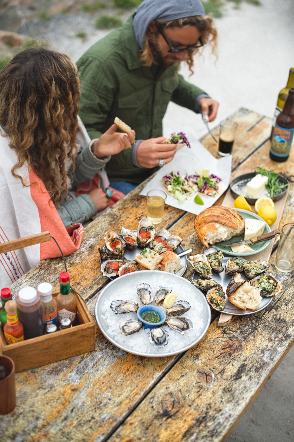 Couple eating oysters and fish tacos at a weathered picnic table on the coast