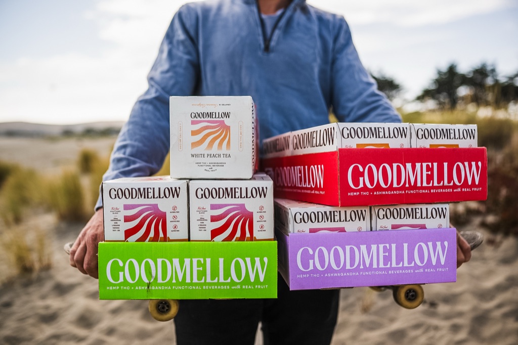 Man carrying colorful cases of Goodmellow functional beverages stacked on skateboards at the beach