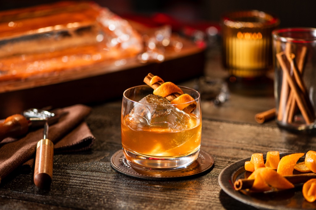 Old fashioned whiskey cocktail with a cinnamon stick and orange twist on a leather coaster in a moody bar