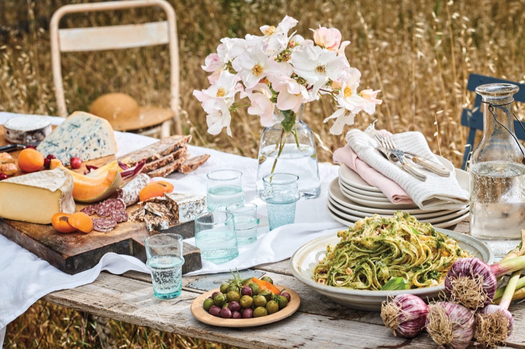 Sunlit outdoor table in a golden field with pesto tagliatelle, a cheese and charcuterie board, and olives