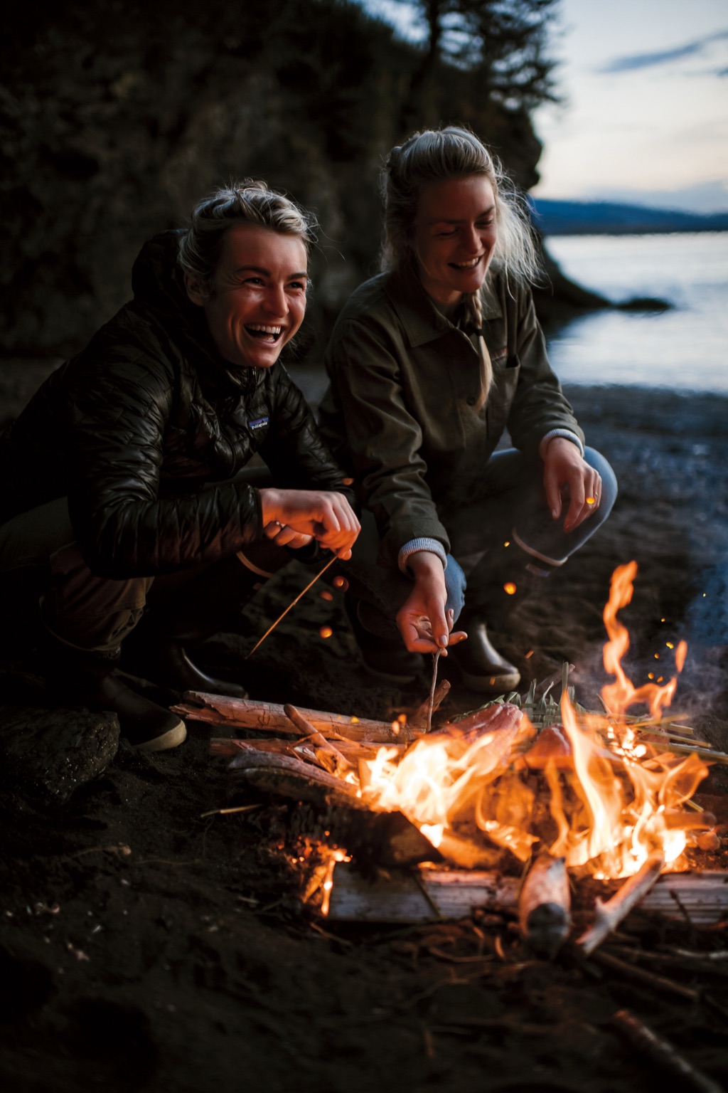The Salmon Sisters laughing while cooking salmon over a driftwood beach campfire at dusk in Alaska
