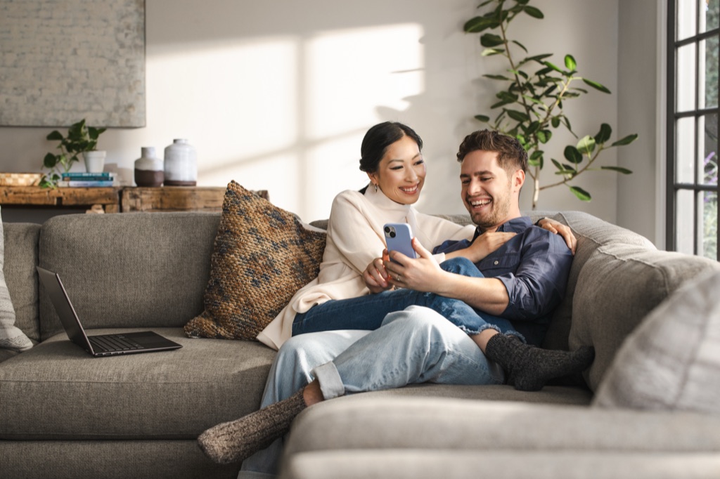 Couple laughing at a phone together on a gray couch in the sun