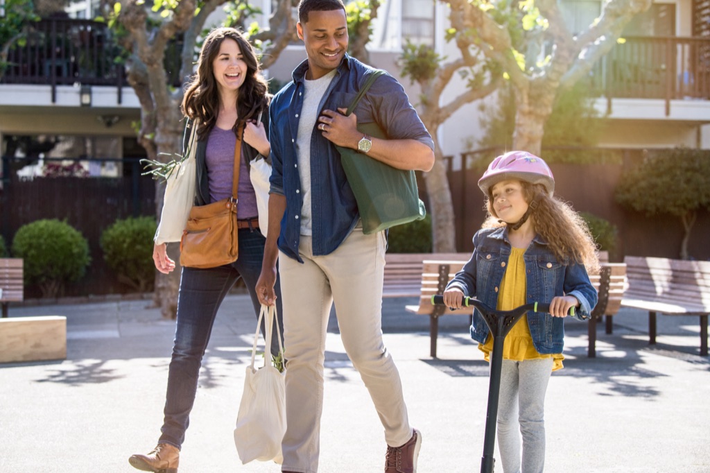 Parents with grocery totes walking beside their daughter riding a scooter in a pink helmet