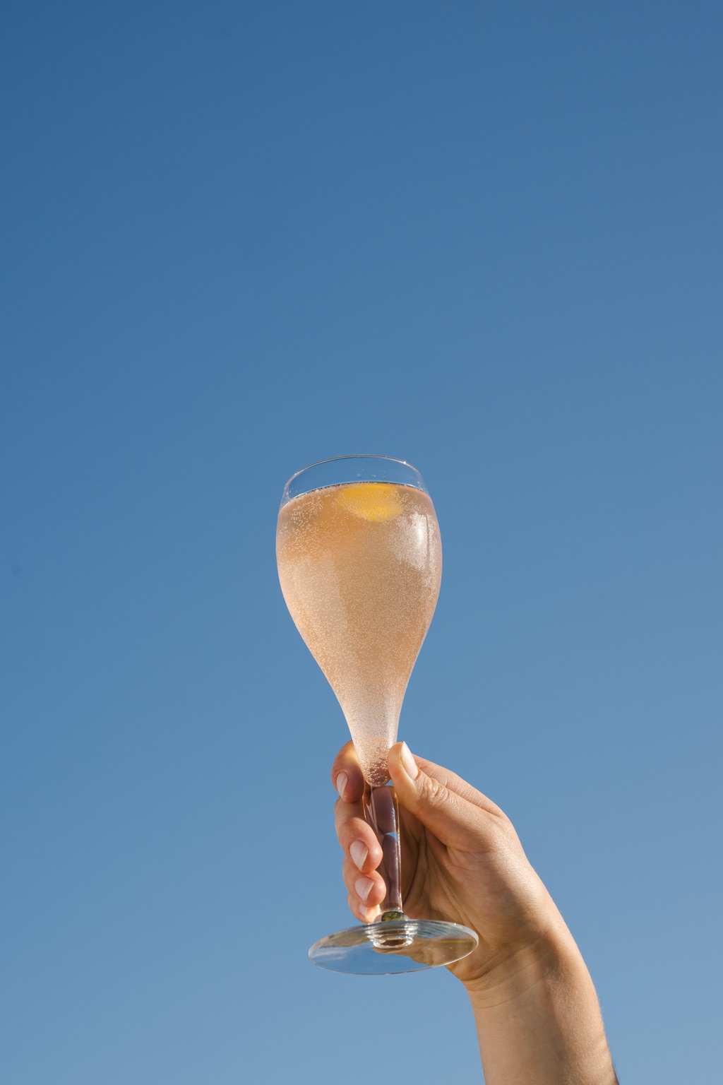 A hand raising a glass of sparkling rose against a clear blue sky, photographed by Dawn Heumann