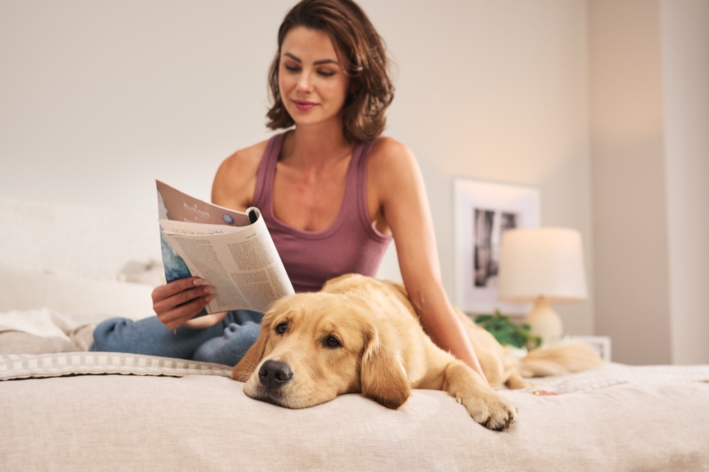 Woman reading a magazine on a bed while a golden retriever rests its head on the duvet