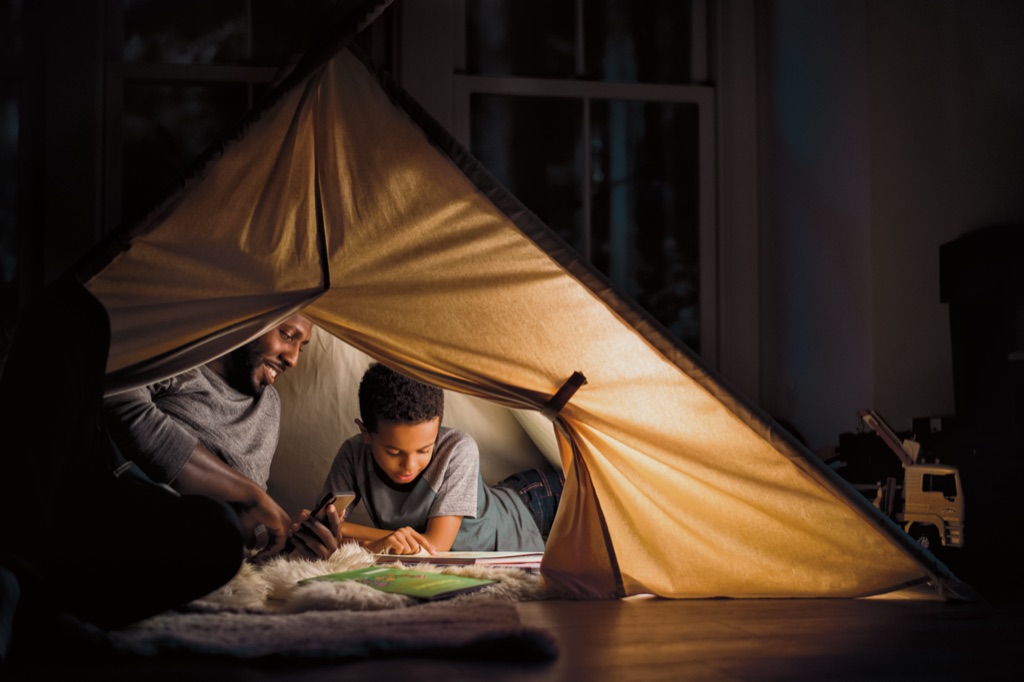 Dad and son reading books inside a glowing lamplit blanket fort at night