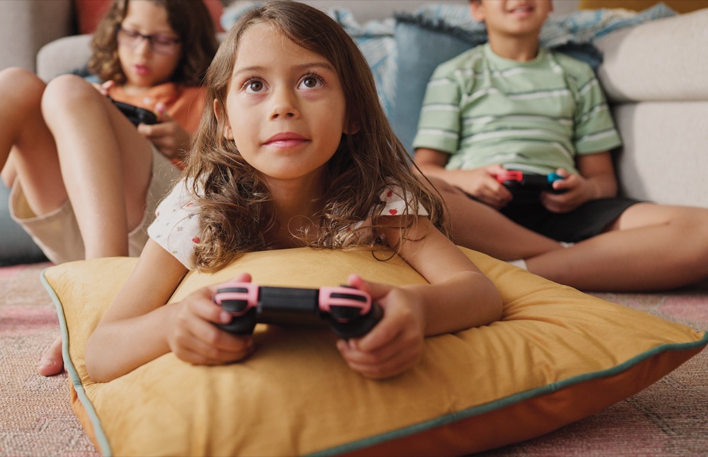 Girl resting on a yellow floor pillow holding a pink game controller with friends behind her