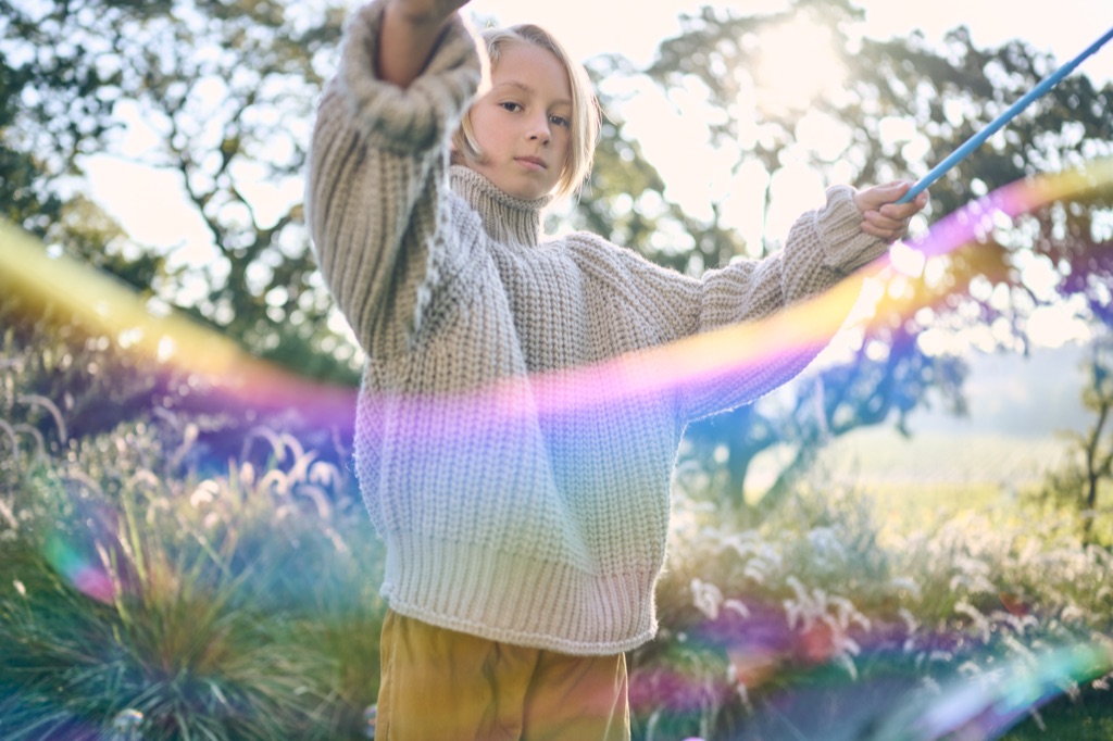 Girl in a cream turtleneck sweater swinging a giant rainbow bubble wand in a sunlit field