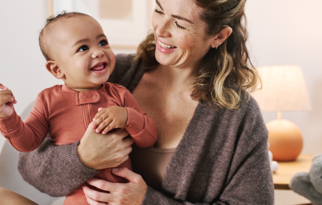 Mom smiling down at the laughing baby in her arms in a warm lamplit nursery