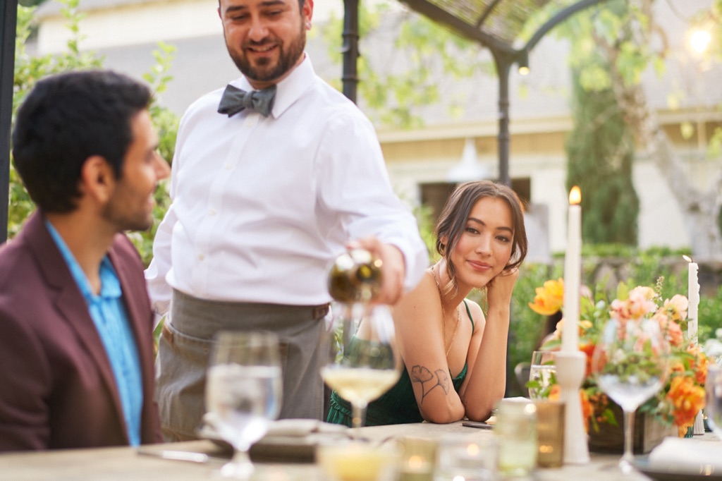 Server in a bow tie pouring white wine at a candlelit garden dinner party in Sonoma County