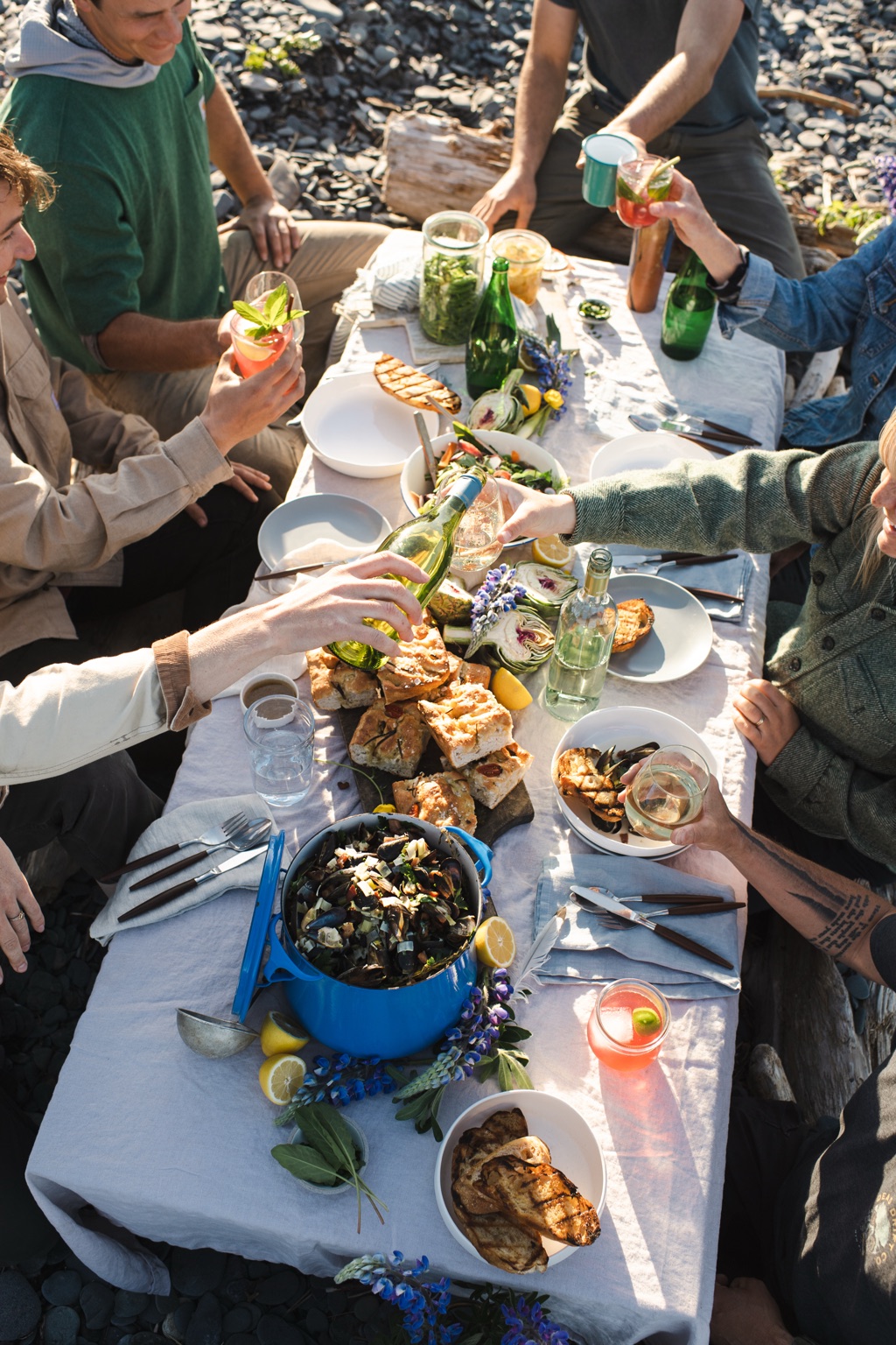Overhead of a driftwood beach table with a pot of mussels, focaccia, and friends pouring white wine