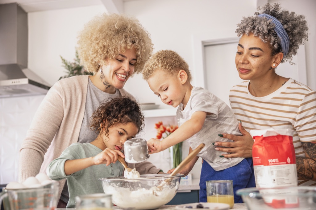 Two moms baking with two young kids scooping flour into a glass bowl