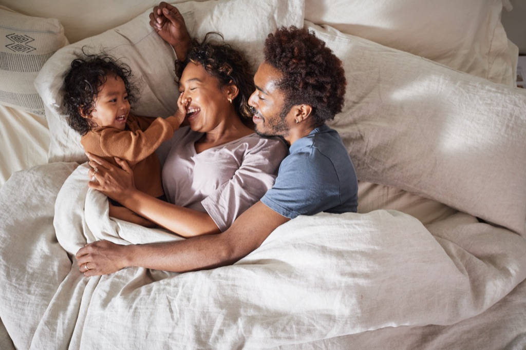 Overhead of a family in bed, a toddler touching his laughing mom's nose while dad hugs them close