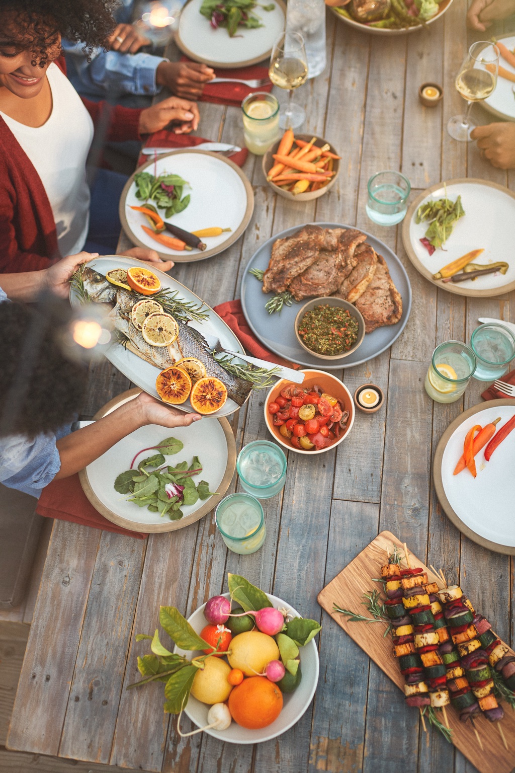 Overhead of an outdoor farm table with a woman serving whole roasted fish, grilled citrus, and vegetable skewers