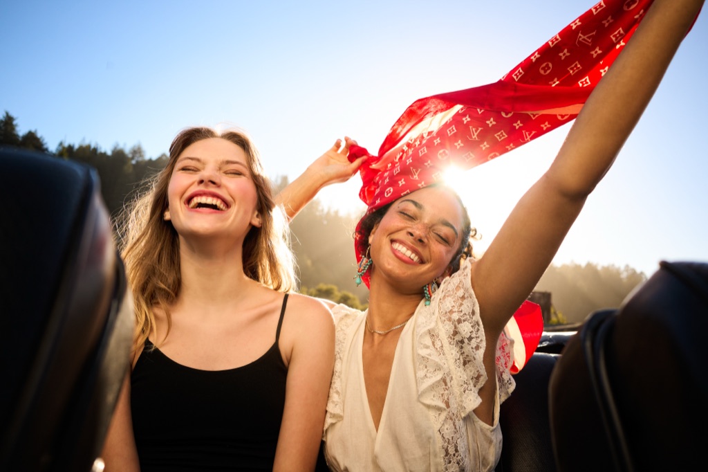 Two young women laughing in the back of a convertible, one waving a red patterned scarf in the sun
