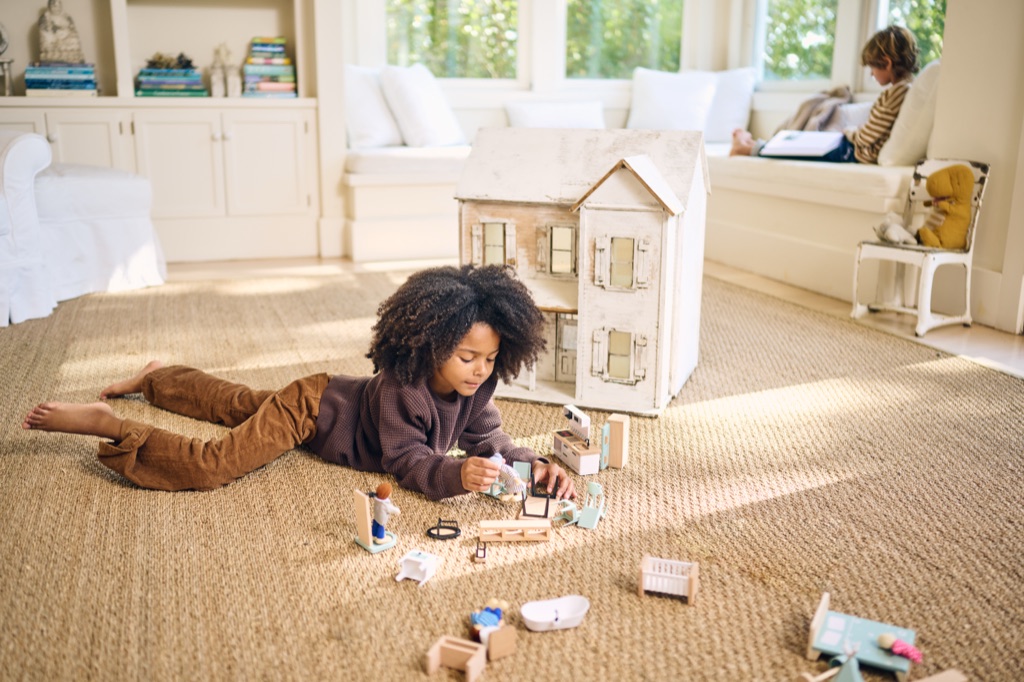 Girl lying on a sisal rug arranging dollhouse furniture while a boy reads on a window seat behind her