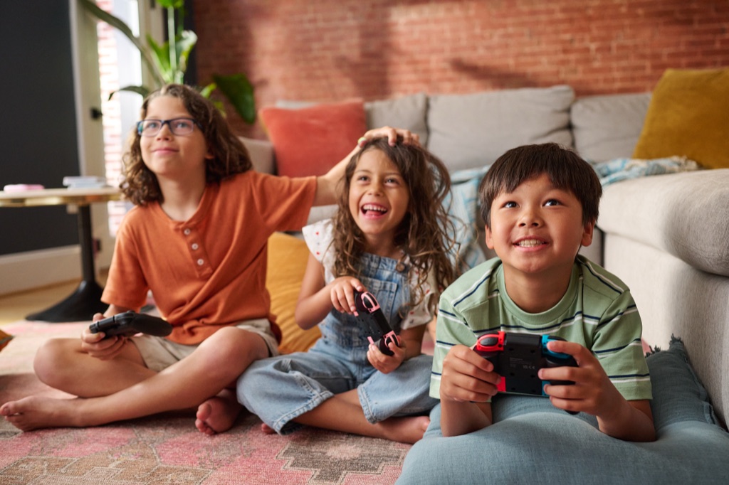 Three kids on a living room floor laughing while playing video games with controllers