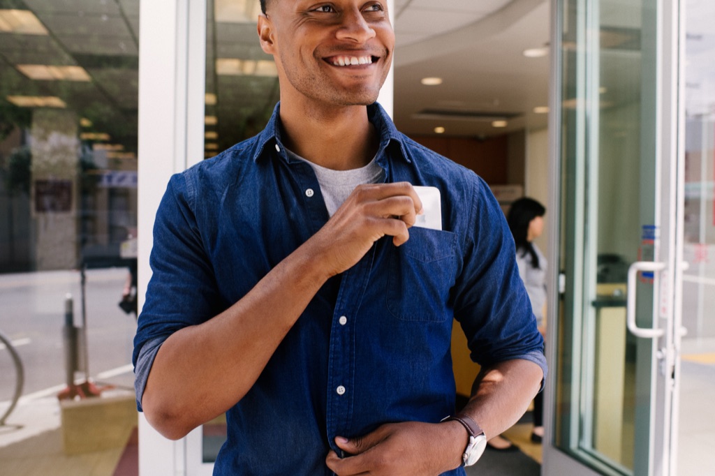 Smiling man in a denim shirt slipping a card into his shirt pocket outside a bank