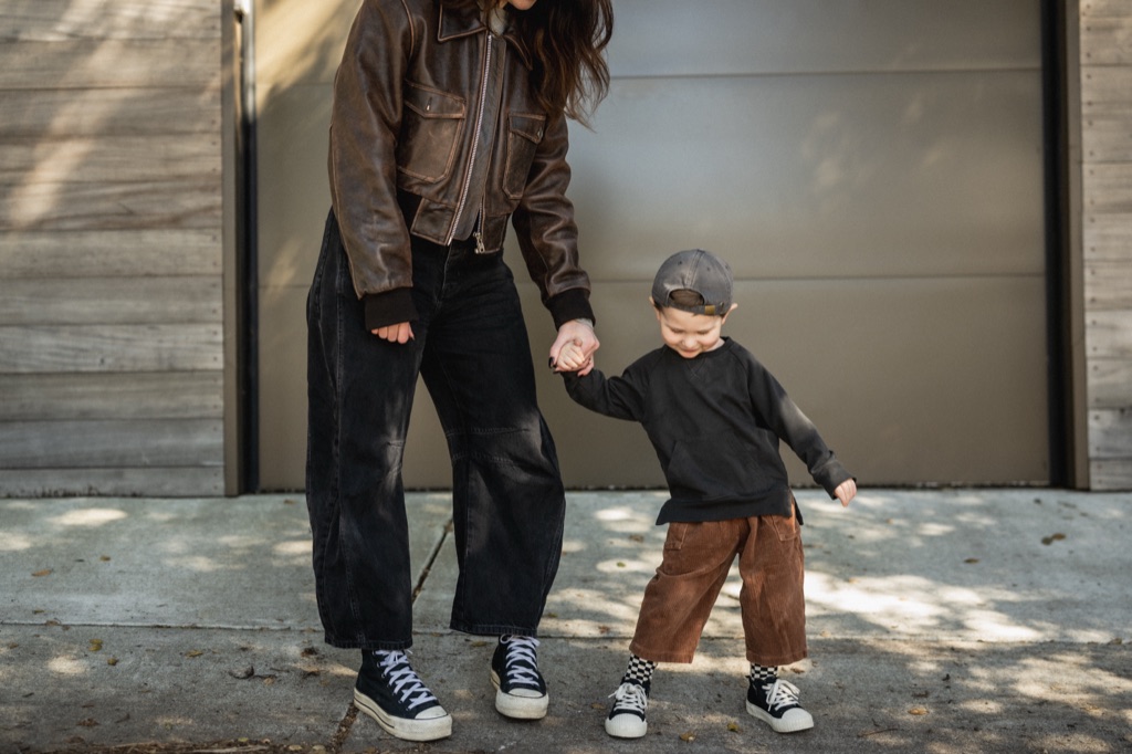 Mom in a leather jacket holding hands with her toddler in a backwards cap on the sidewalk