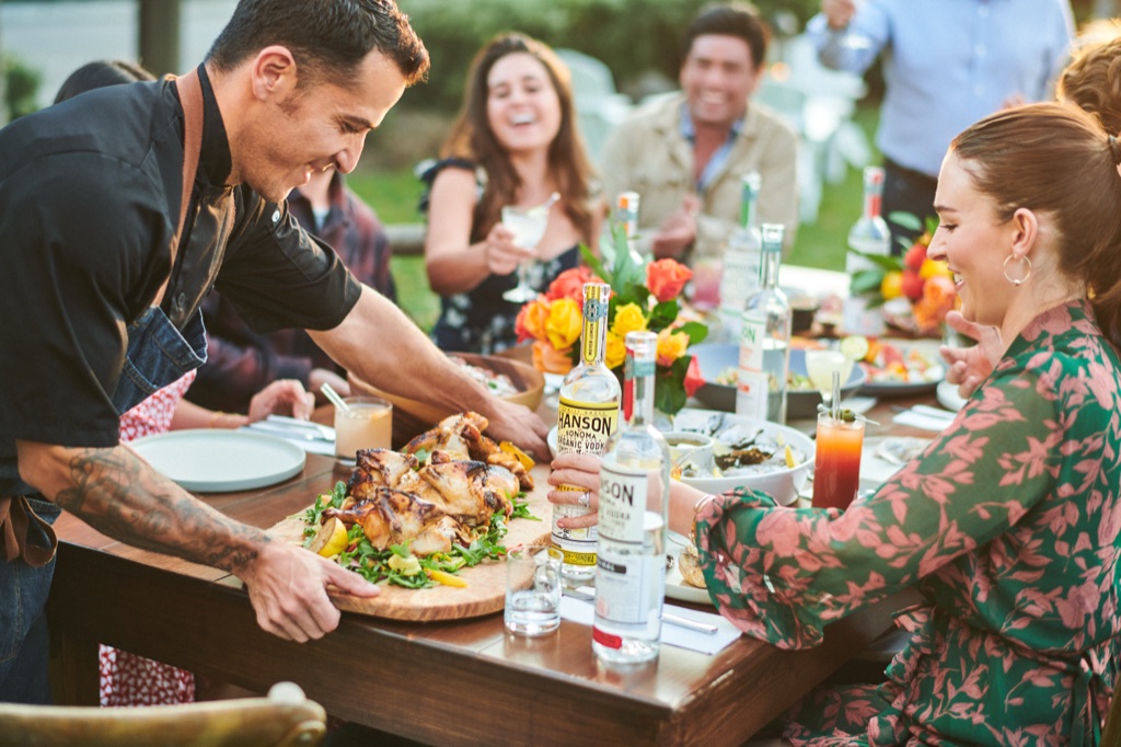 Chef setting a platter of grilled chicken on an outdoor dinner table set with Hanson of Sonoma organic vodka