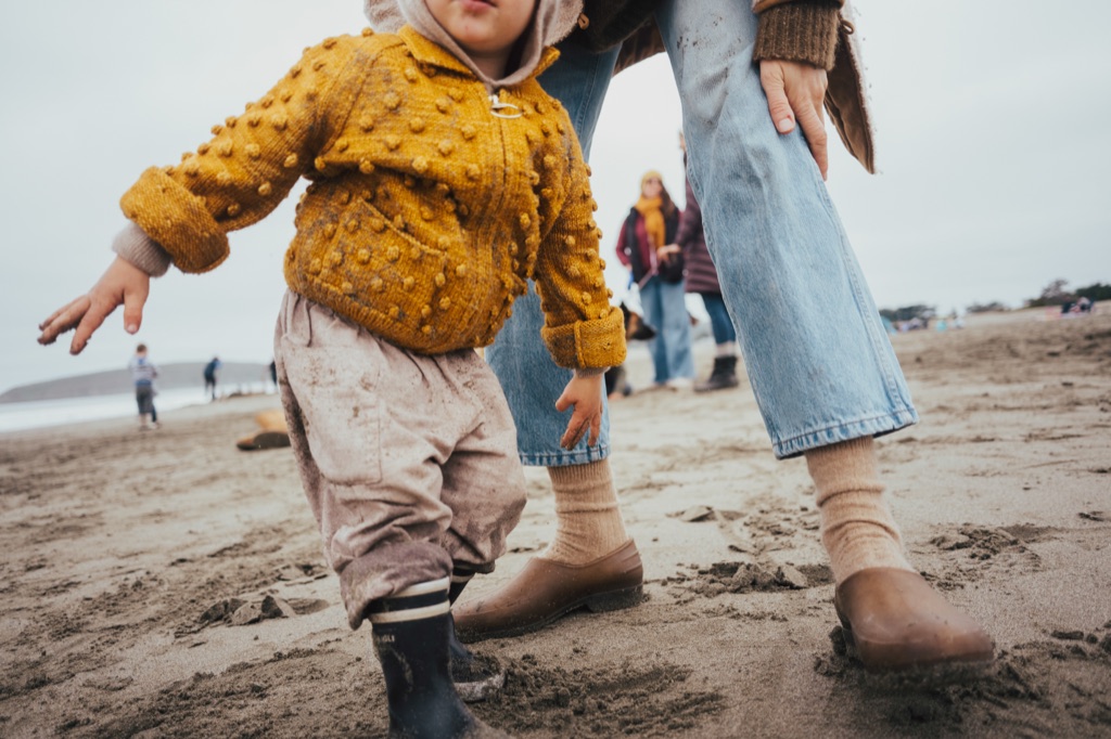 Toddler in a popcorn knit sweater and rain boots standing on an overcast beach