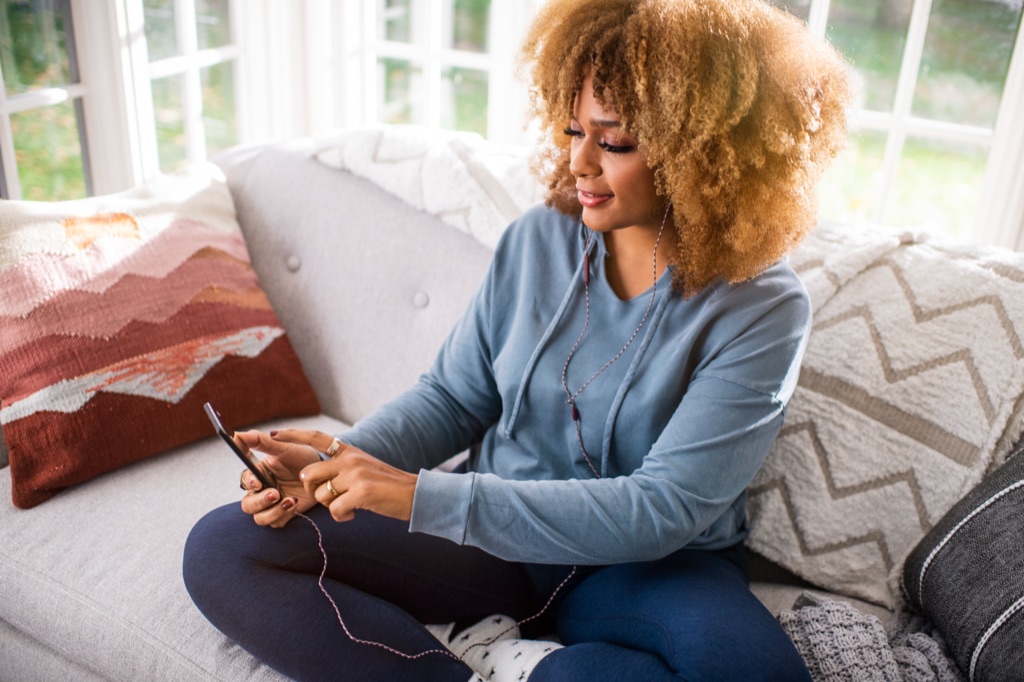 Woman with golden curls sitting cross legged on a sunroom couch listening with corded earbuds and scrolling her phone