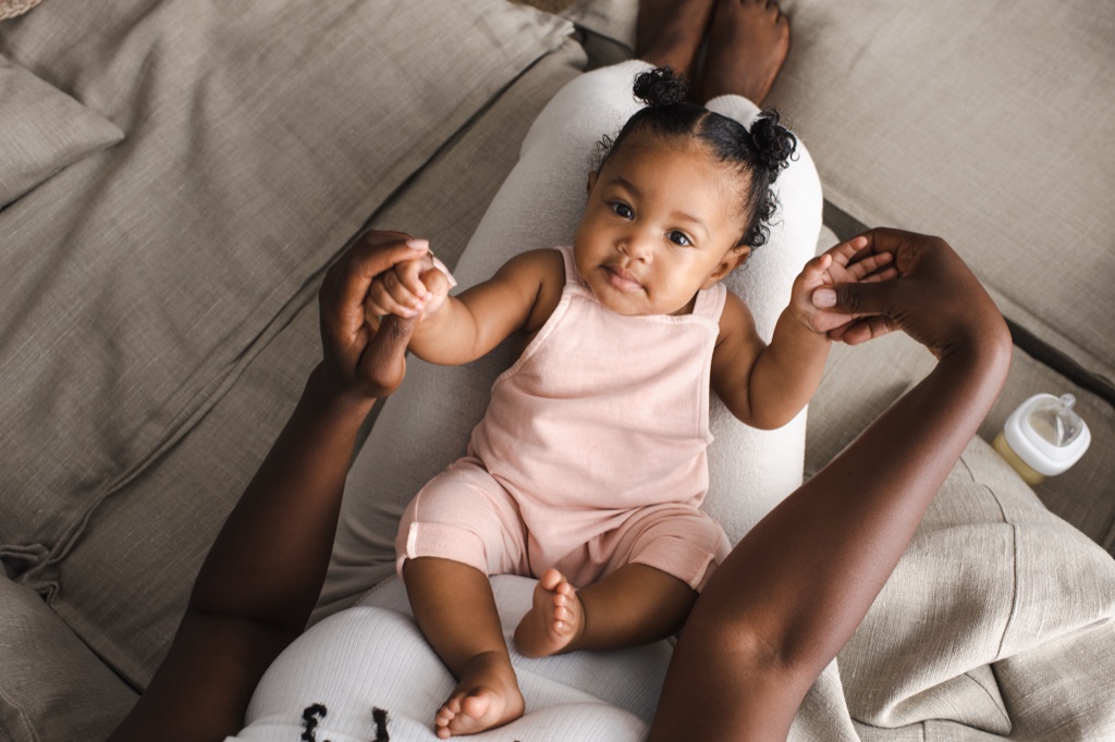 Baby girl with curly pigtail buns in a pink romper resting on a nursing pillow and holding a parent's fingers