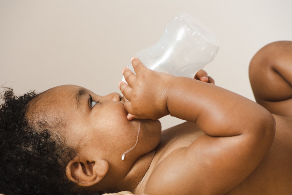 Baby lying back drinking from a bottle held in its own hands, milk dribbling down one cheek